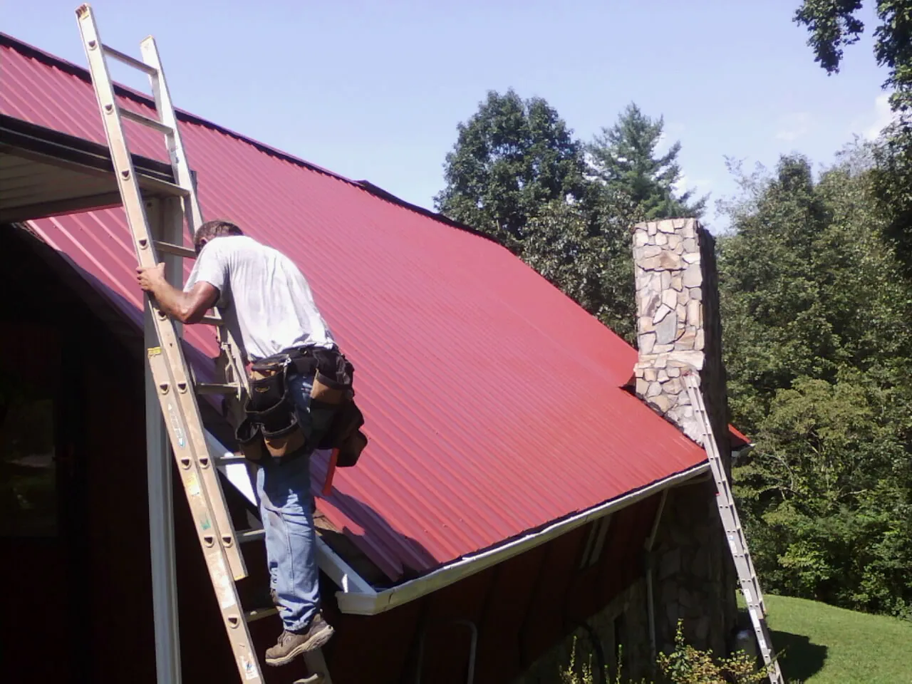 Roofer on a ladder beside a red metal roof and stone chimney — Soffit & Fascia Repair by Roofing Brothers in Pilot